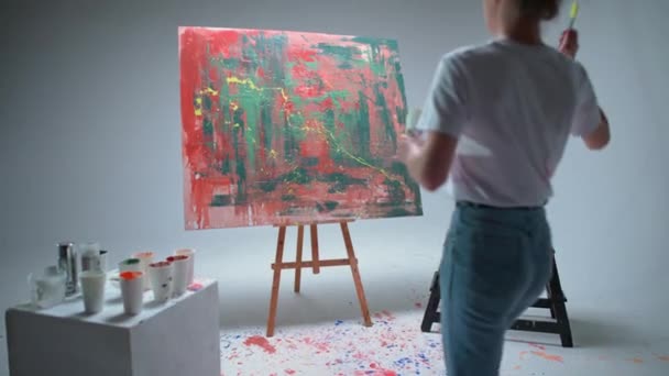 Handheld, ženský umělec kreslí štětcem na velké plátno v bílém pokoji, talentovaný umělec kreslí barevné červené abstrakce, moderní umění v olejomalbě. — Stock video