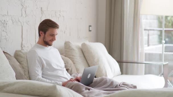 Homem caucasiano alegre senta-se em casa no sofá e trabalha usando um laptop, gerente financeiro em um trabalho remoto durante o auto-isolamento em quarentena, um homem faz um bom negócio, emoções positivas, câmera lenta — Vídeo de Stock