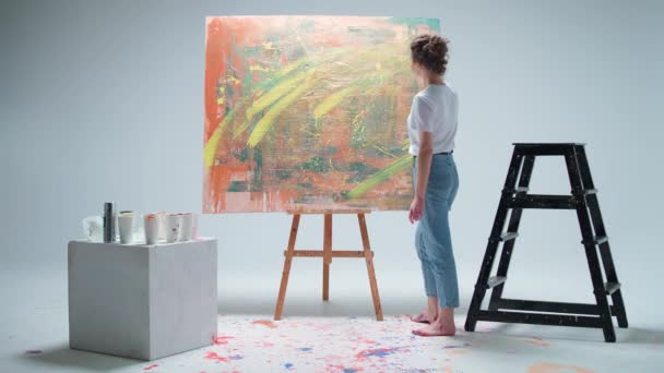 Arte moderno, joven artista femenina dibuja con pintura en aerosol en un gran lienzo en una habitación blanca, un artista talentoso dibuja una abstracción de color, vista posterior. — Vídeos de Stock