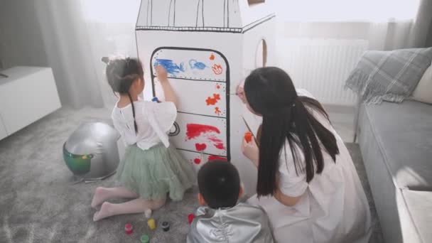 子供を持つアジアの女性は自宅のリビングルームで遊ぶ、彼女の母親と妹と一緒に床に座って宇宙飛行士の衣装の男の子、子供たちは段ボールモデルに母親のペイントと一緒に — ストック動画