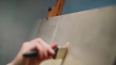 İşteki yaratıcı kişi, yetenekli bir yetişkin adam ressam bej yağlı boya ile tuvali kaplıyor, arka planı çiziyor, ellerin yakın görüntüsü, 4k yavaş çekim.