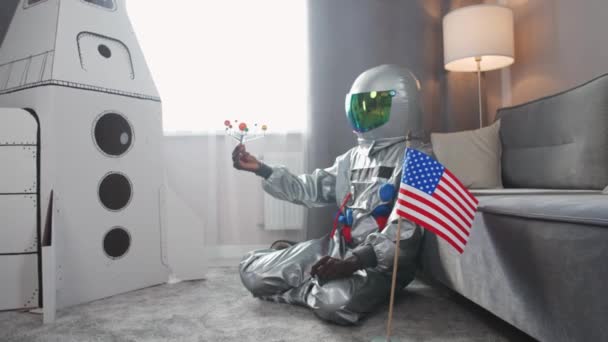 Африканский мужчина в американском костюме астронавта сидит на полу в гостиной дома и играет с игрушечной моделью солнечной системы, человек на самоизоляции, флаг США на переднем плане, 4k — стоковое видео