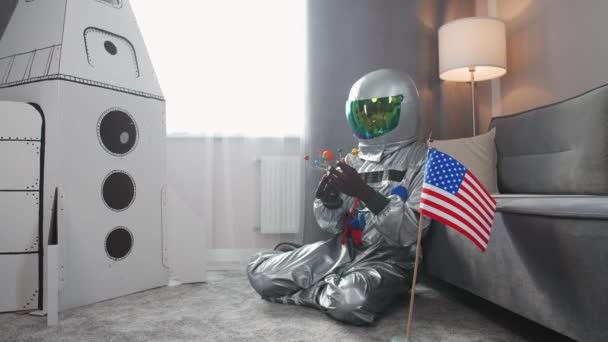 Afroamerikanischer Mann im Astronautenkostüm sitzt zu Hause im Wohnzimmer auf dem Boden und spielt mit einem Spielzeugmodell des Sonnensystems, Mann in Selbstisolation, im Vordergrund die Flagge der USA, 4k — Stockvideo