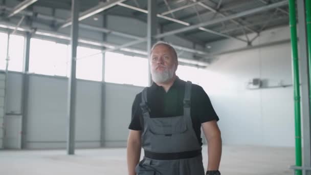 Retrato de trabajador de pelo gris camina a través del almacén, un ingeniero de trabajo camina a través del territorio de un almacén logístico vacío, 4k cámara lenta. — Vídeo de stock