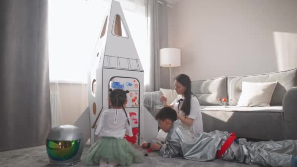 Aziatische vrouw met haar kinderen spelen in de woonkamer thuis, een jongen in een astronaut kostuum liggend op de vloer, kinderen samen met hun moeder verf op een kartonnen model van een ruimteschip met verf. — Stockvideo