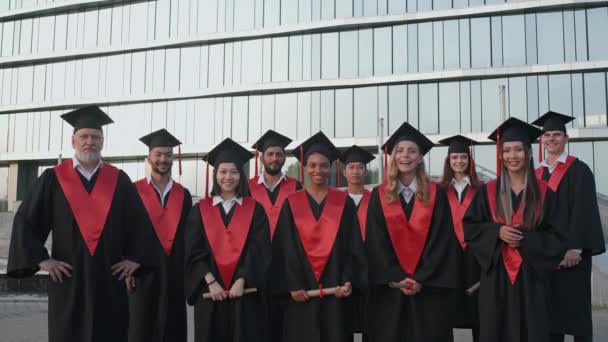 Los graduados universitarios se regocijan al final de sus estudios, las personas de diferentes edades y razas en la túnica de los graduados que están de pie cerca de la universidad moderna, los graduados alegres miran a la — Vídeo de stock