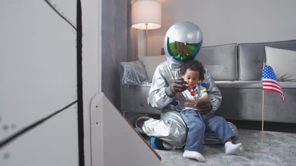 Афроамериканец отец и сын играют в гостиной у себя дома, мужчина в костюме американского астронавта сидит на полу со своим сыном, мальчик играет с игрушечной моделью солнечной системы, изучает — стоковое видео