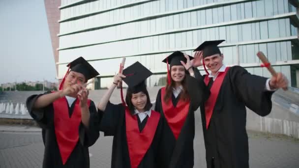 Les diplômés universitaires se réjouissent à la fin de leurs études, les gens de différentes races en robe de diplômés ils se tiennent près de l'université moderne et jettent des chapeaux dans l'air, les diplômés joyeux regardent — Video