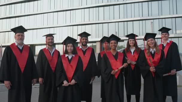 Graduados universitarios se regocijan al final de sus estudios, personas de diferentes edades y razas en la túnica de los graduados que están de pie cerca de la universidad moderna, graduados alegres mirar a la cámara — Vídeos de Stock