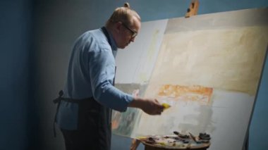 Sanatçılar atölyesi, yaratıcı kişi, yetenekli bir yetişkin adam ressam bej rengi boya ile tuvali kaplıyor, modern soyut bir resim çiziyor, 4k yavaş çekim..