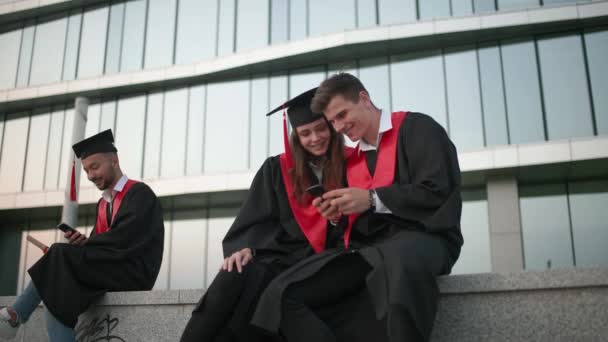 A középiskolai végzősök örvendeznek tanulmányaik végén, a diplomás köpenyes fiatalok az egyetem közelében ülnek és okostelefonokat használnak, képeket néznek, pozitív érzelmeket. — Stock videók