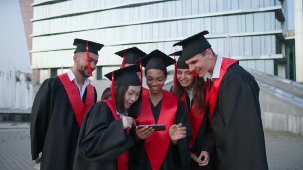 Graduação em uma universidade internacional, jovens em vestes de pós-graduação em pé perto da universidade e visualização de fotos no smartphone, emoções positivas. — Vídeo de Stock