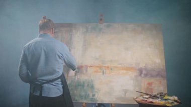 Sanatçılar atölyesi, yaratıcı kişi, yetenekli bir yetişkin adam ressam bej rengi boya ile tuvali kaplıyor, modern soyut bir resim çiziyor, arka plan, 4k yavaş çekim..