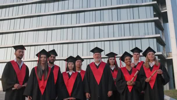 Los graduados universitarios internacionales se regocijan al final de sus estudios, las personas de diferentes edades y razas en la túnica de los graduados que están de pie cerca de la universidad moderna, graduados alegres miran — Vídeo de stock