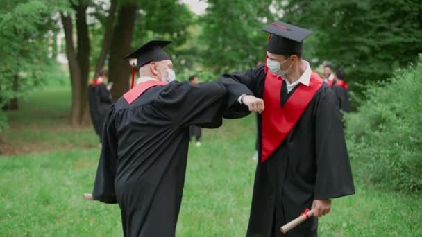 Internationellt universitet, examen, lärare gratulerar mannen student för att få sitt diplom, lärare och student i medicinska masker hälsa varandra med armbågar, 4k slow motion. — Stockvideo