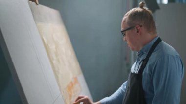 Sanatçılar atölyesi, yaratıcı kişi, yetenekli bir yetişkin adam ressam bej rengi boya ile tuvali kaplıyor, modern soyut bir resim çiziyor, 4k yavaş çekim..