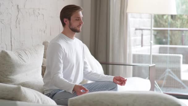 Mindfulness och lugn stämning, ung man sitter på en soffa i vardagsrummet och mediterar, en lugn stämning, koppla av i ett vitt rum fyllt med ljus. — Stockvideo