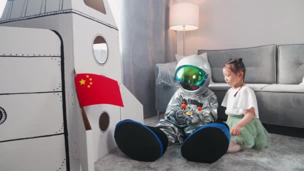 Azjatyckie dzieci bawią się w salonie w domu, chłopiec w kostiumie astronauty siedzący na podłodze ze swoją siostrą, dzieci bawiące się zabawkowym modelem układu słonecznego, 4k zwolnione tempo. — Wideo stockowe
