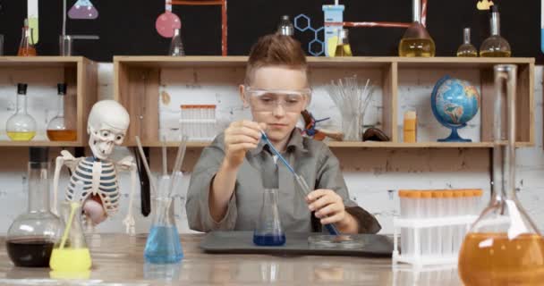 Laboratoriumervaring in een scheikundeles, jongen in beschermende bril giet een blauwe vloeistof in een reageerbuis, modern onderwijs op school. — Stockvideo