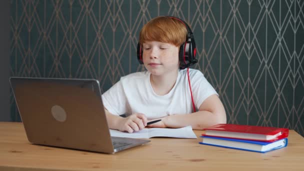 Distans lärande, rödhårig tonåring pojke sitter på bordet i vardagsrummet och använder en bärbar dator för att studera i självisolering hemma, hemundervisning online genom videokall, studera under en pandemi i — Stockvideo
