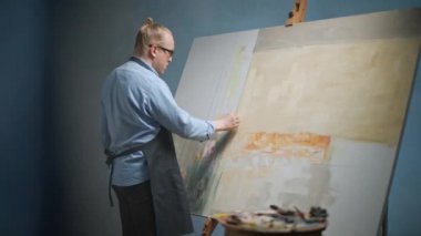 İşteki yaratıcı kişinin arka planında, yetenekli bir erkek sanatçı büyük bir tuval üzerine soyut bir resim çizer, bej yağlı boyayla tuvali kaplar..