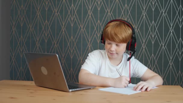 Distance learning, red-haired teen boy sitting on the table in living room and uses a laptop to study in self-isolation at home, homeschooling online by videocall, studying during a pandemic in — Stock Video