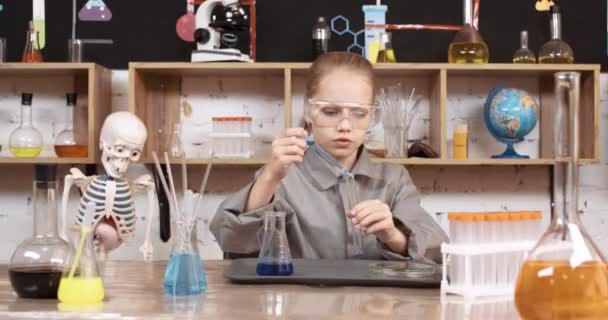 Laboratóriumi tapasztalat egy kémia órán, egy védőszemüveges lány kék folyadékot önt egy kémcsőbe, modern iskolai oktatás. — Stock videók