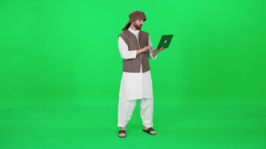 Geleneksel giysiler içinde Arap görünümlü bir adam bir krom anahtarın arka planında dururken internette sörf yapmak için dizüstü bilgisayar kullanır..