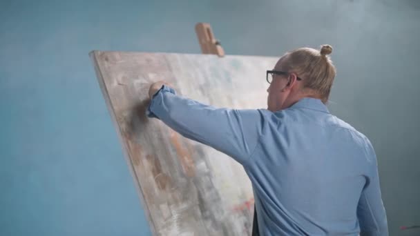 HANDHELD, творча людина в синій сорочці і окулярах на роботі, талановитий дорослий чоловік художник малює абстрактну картину на великому полотні . — стокове відео