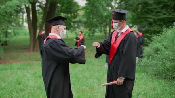 Internationale universiteit, diploma-uitreiking, leraar feliciteert de man student met het ontvangen van haar diploma, leraar en student in medische maskers handen schudden, handdruk vuist hobbel, 4k slow motion. — Stockvideo