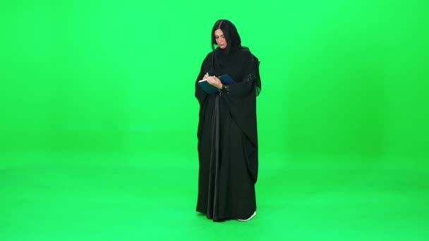 Femme d'apparence arabe en vêtements traditionnels écrit dans un carnet tandis que se dresse sur le fond d'un chromakey. — Video