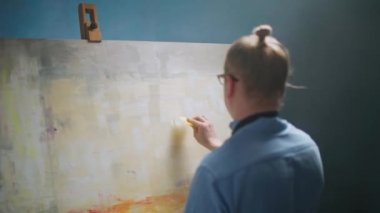 Sanat stüdyosu, mavi gömlekli ve gözlüklü yaratıcı kişi, yetenekli yetişkin bir erkek sanatçı, büyük bir tuvale soyut bir resim çizer..