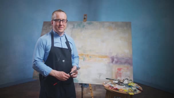 Portret van een volwassen mannelijke kunstenaar tegen de achtergrond van een groot doek, een man staat in zijn atelier en kijkt naar de camera, creatieve persoon in een blauw shirt en bril op het werk. — Stockvideo