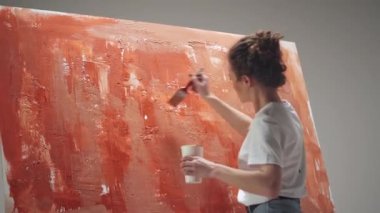 Genç bir kadın sanatçı, beyaz bir odada büyük bir tuvale fırçayla resim çizer. Yetenekli bir sanatçı, kırmızı bir soyutlama çizer..