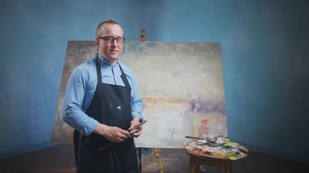 Zpomalený portrét dospělého muže, umělce stojícího na pozadí velkého plátna, muže stojícího ve svém ateliéru a dívajícího se na kameru, kreativního člověka v modré košili a brýlích — Stock video