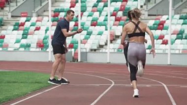 Stadyumda koşucular antrenman yapıyor, iki sportif kadın kısa mesafe koşusunda yarışıyor, koç zamanı kontrol ediyor..
