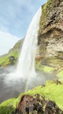 İzlanda şelalesinin görkemli manzarası, İzlanda doğası ve dünya dokusunun huzur manzarası, su ve gökkuşağı etkisinin akışı, yeşil kayalık arazi arka planı.