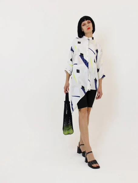 Kısa siyah kısa kısa saçlı, geometrik desenli asimetrik gömlekli siyah ip torbalı ve çantasının içinde tenis açık yeşil topları olan genç bir kız model. Kıyafet kataloğu için çekim