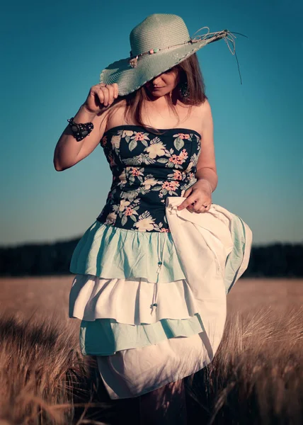 Korseli bir kızın fotoğrafı, beyaz ve mavi tüylü bir etek ve geniş kenarlı buğday kulaklı mavi bir şapka. Buğdaydan altın kulaklar ve tarlada güzel, genç bir kız.