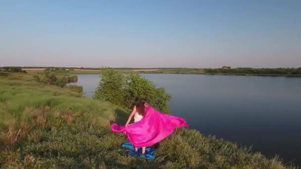 Dziewczyna z luźnymi włosami w sukience stoi na różowej tkaninie na wysokim brzegu jeziora. Widok z drona. — Wideo stockowe