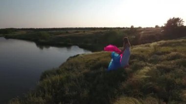 Elbiseli, bol saçlı bir kız gölün yüksek kıyısında pembe ve mavi bir bezle oynuyor. İnsansız hava aracının dairesel görüntüsü.