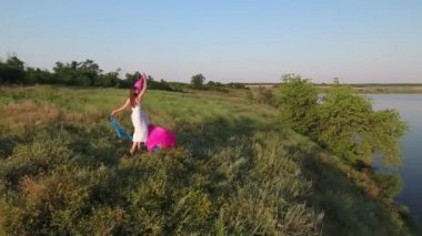 Elbiseli, bol saçlı bir kız gölün yüksek kıyısında pembe ve mavi bir bezle oynuyor. Bir İHA görüntüsü.