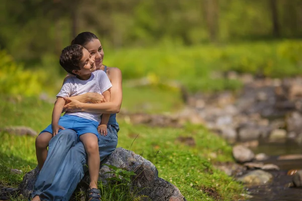 Молодая мать с сыном сидит на камне возле ручья в лесу — стоковое фото