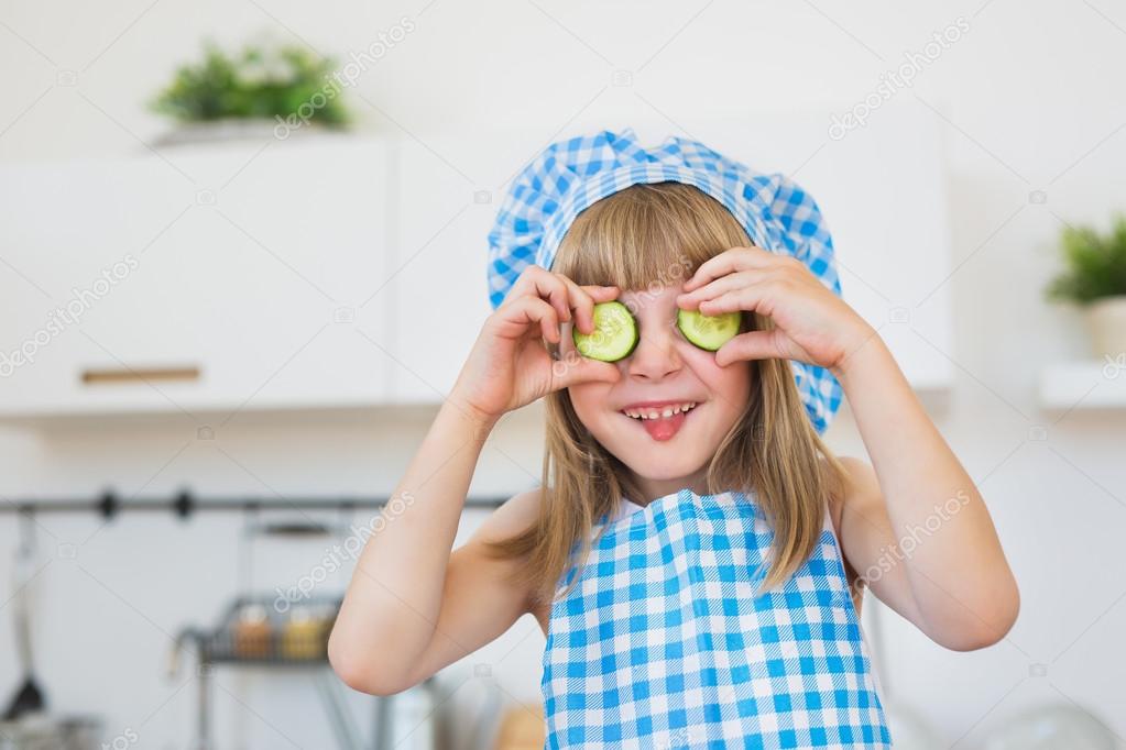 Funny little girl in cook clothes holds a slices of a cucumber on an eyes and puts out a tongue