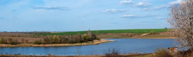Güneşli bir günde gölün yatay manzarası. İlkbaharda ağaç beyaz çiçeklerle açar. Gölün üstünde yeşil bir buğday tarlası var. ve mavi gökyüzü.