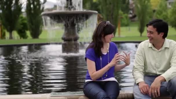 A shot of a young couple conversing with each other in front of a water fountain. — Stock Video