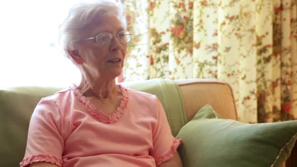 Una foto estática de una anciana con una blusa rosa hablando en un sofá amarillo. Tiene pelo gris y gafas circulares. El sofá tiene almohadas verdes . — Vídeos de Stock