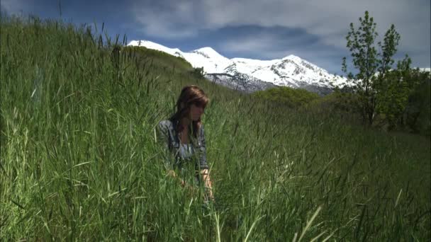 Fotografía en cámara lenta de una joven sentada en la hierba alta con montañas detrás de ella . — Vídeos de Stock