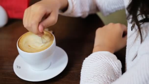 Dama en un pulloverholding blanco una cucharadita y revolviendo preparado delicioso café caliente con leche en una cafetería blanca cupat . — Vídeo de stock