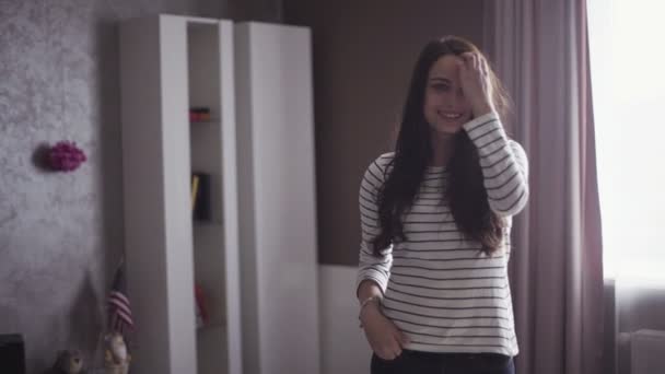 Slow motion portrait of a young cheeful woman who stands in a living room and touches her hair. — Stockvideo